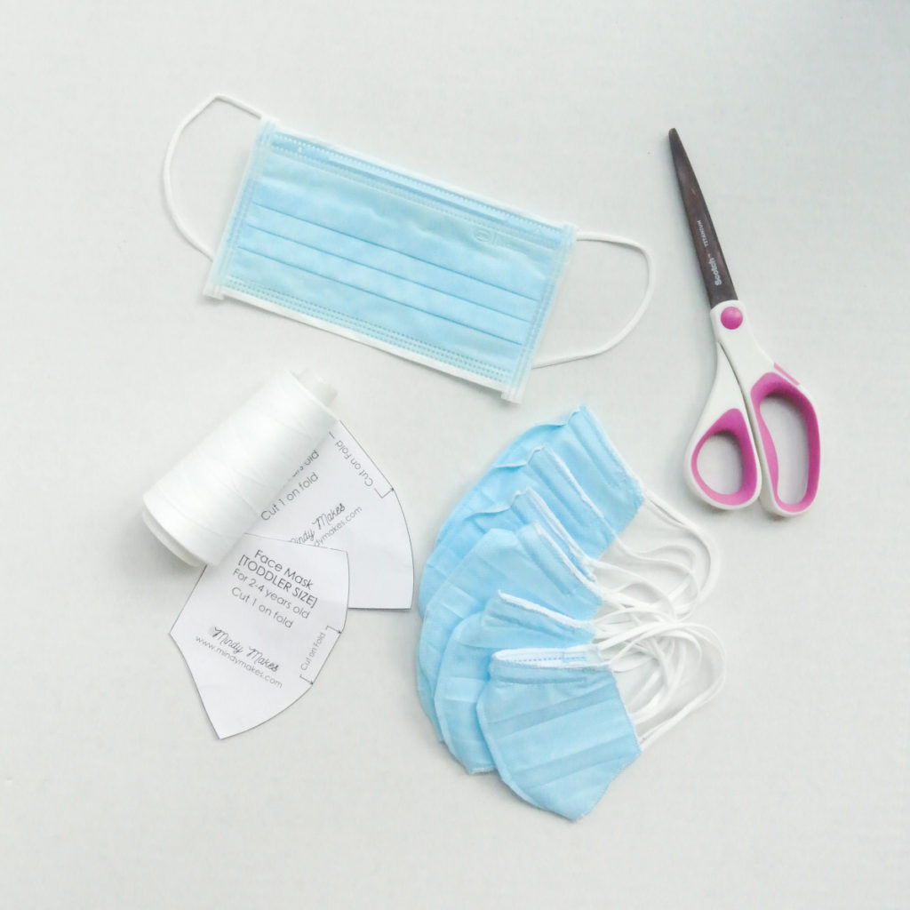 Image shows adult surgical mask, scissors, face mask pattern, a spool of white thread, and 6 finished toddler sized surgical mask